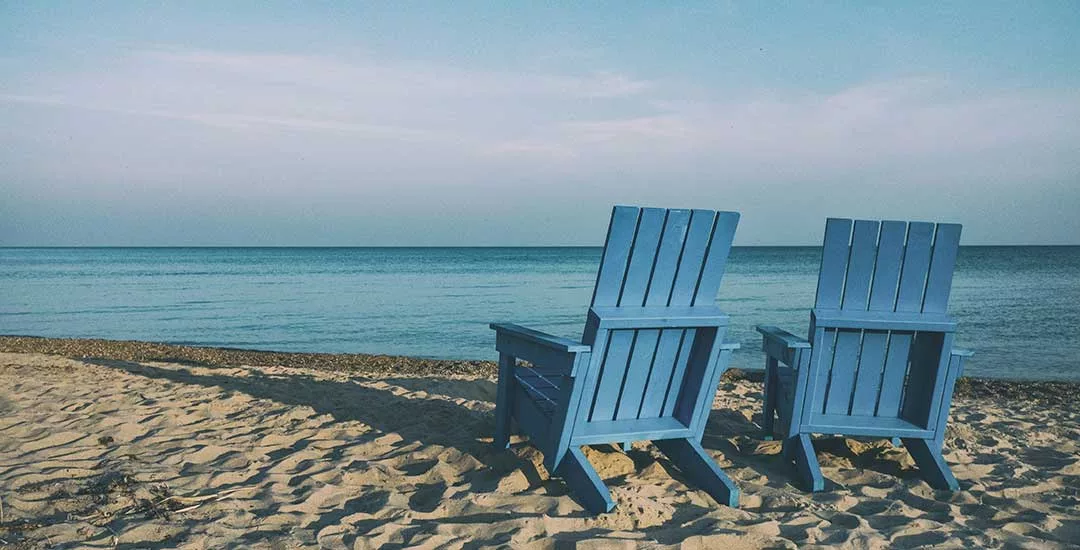 two empty blue wooden deck chairs sitting on a sandy beach facing a very calm blue sea