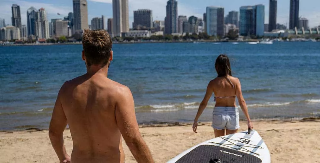 young couple carrying a paddle board to the water with the skyline of a city in the background