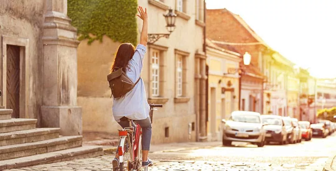back of a woman in a blue shirt and pants riding on a red bike down a cobbled street with her right hand raised in the air.