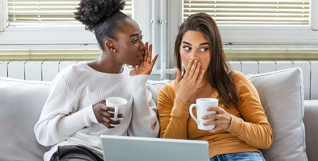 two young women sitting on a couch holding cups of tea with lap top rested on coffee table in front of them looked shocked