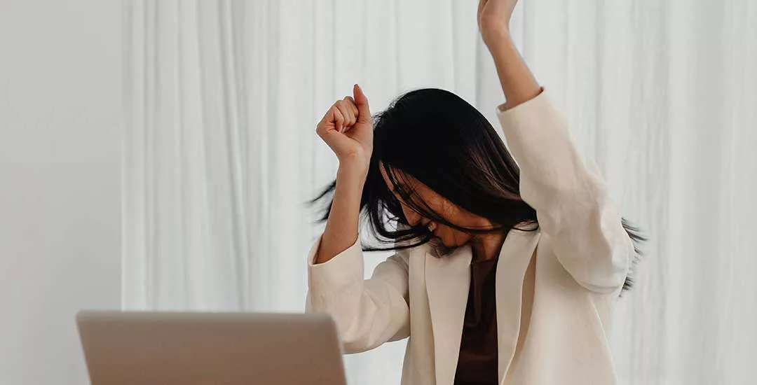 young woman sitting in front of a computer with her hands raised in the air in excitement