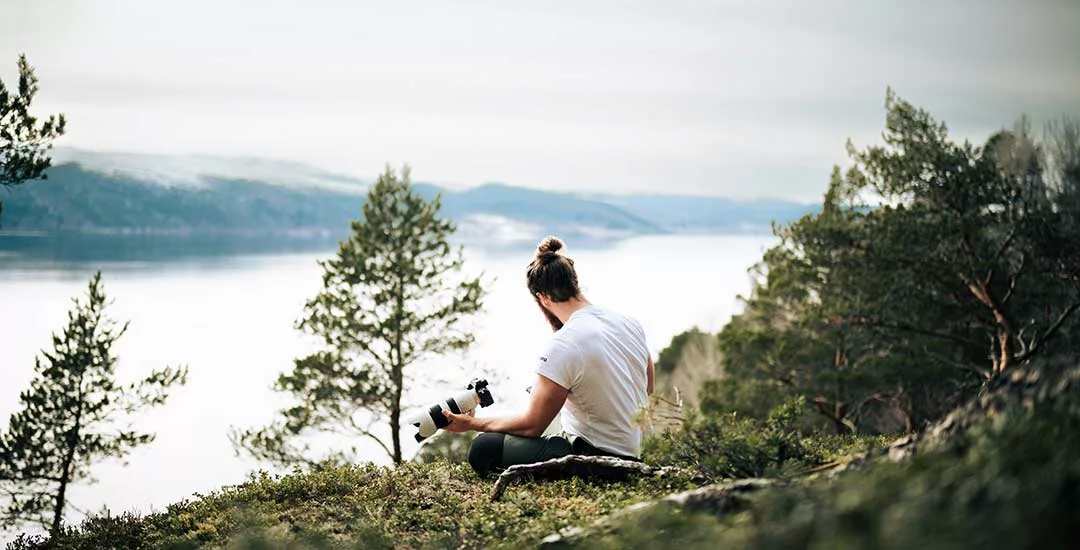 man sitting at the top of a hill range looking down onto a lake with a camera in one hand
