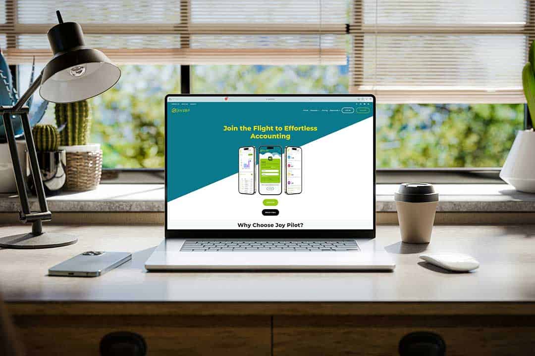 front on view of a laptop sitting on a tidy desk with a view of trees through a wall of windows where blinds are partially drawn, disposable coffee cup to the right, lamp to the left and the screen displaying the home page of www.joypilot.co website